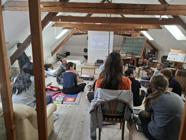 Účastníci sedí na podlaze a židlích a sledují projekci v podkrovním prostoru, pohled přes dřevěný rám